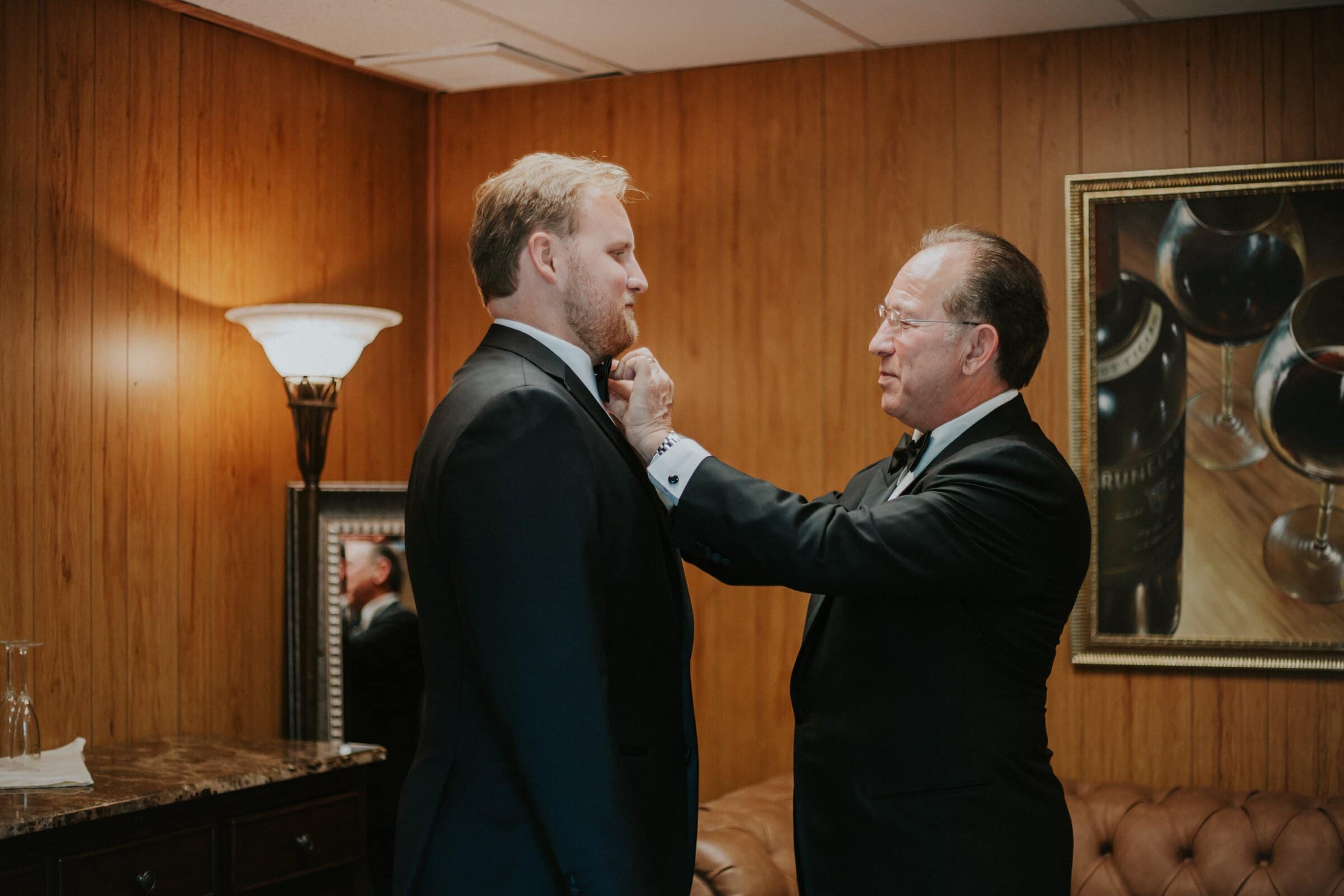 An older man straightening the groom's tie in a wood-paneled room