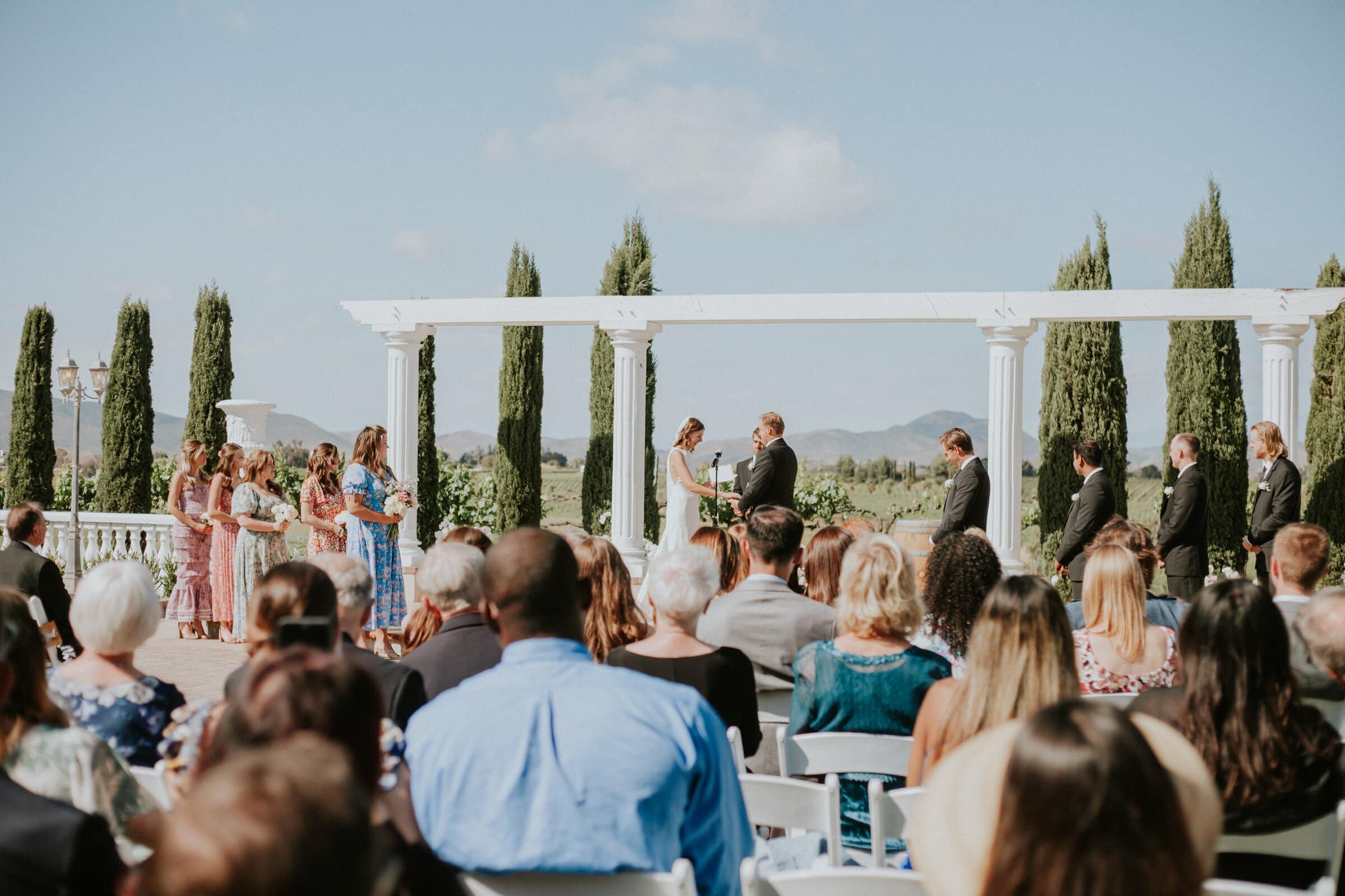 Guests seated in the sun watching the ceremony under the white colonnade