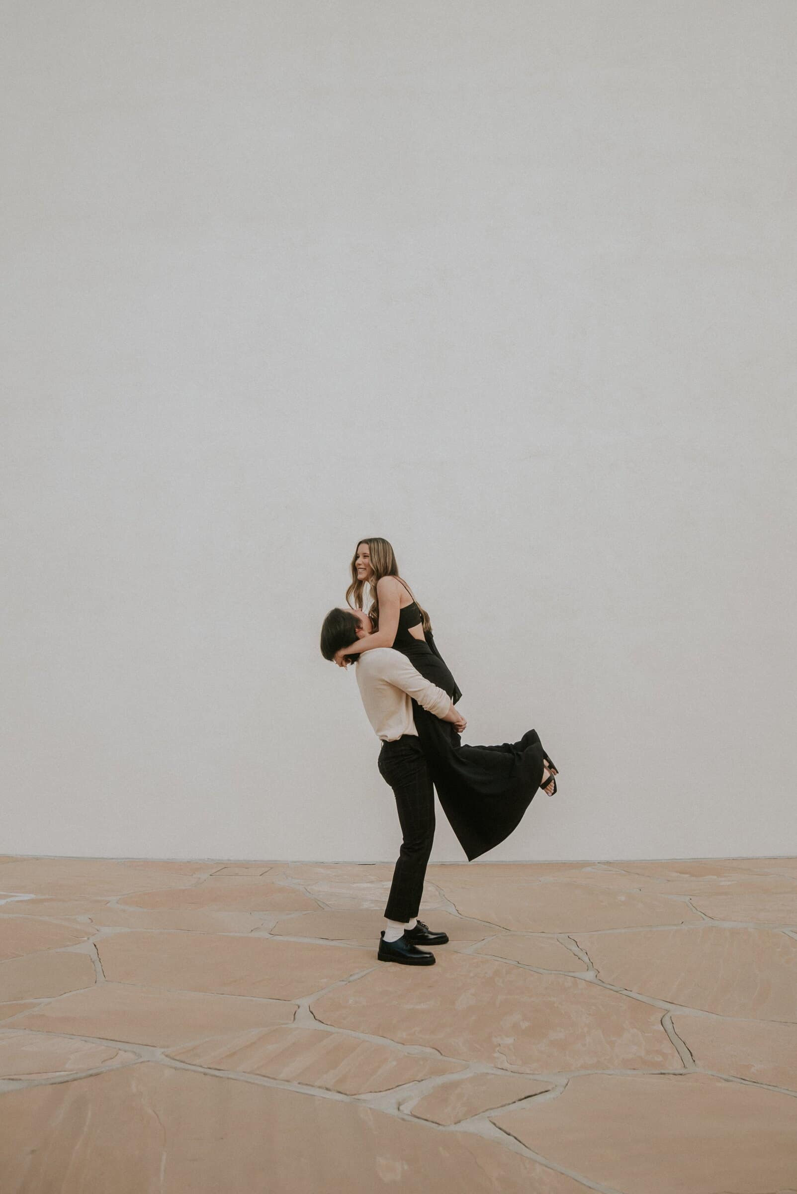 Cameron lifting Amanda off her feet against a bare white wall