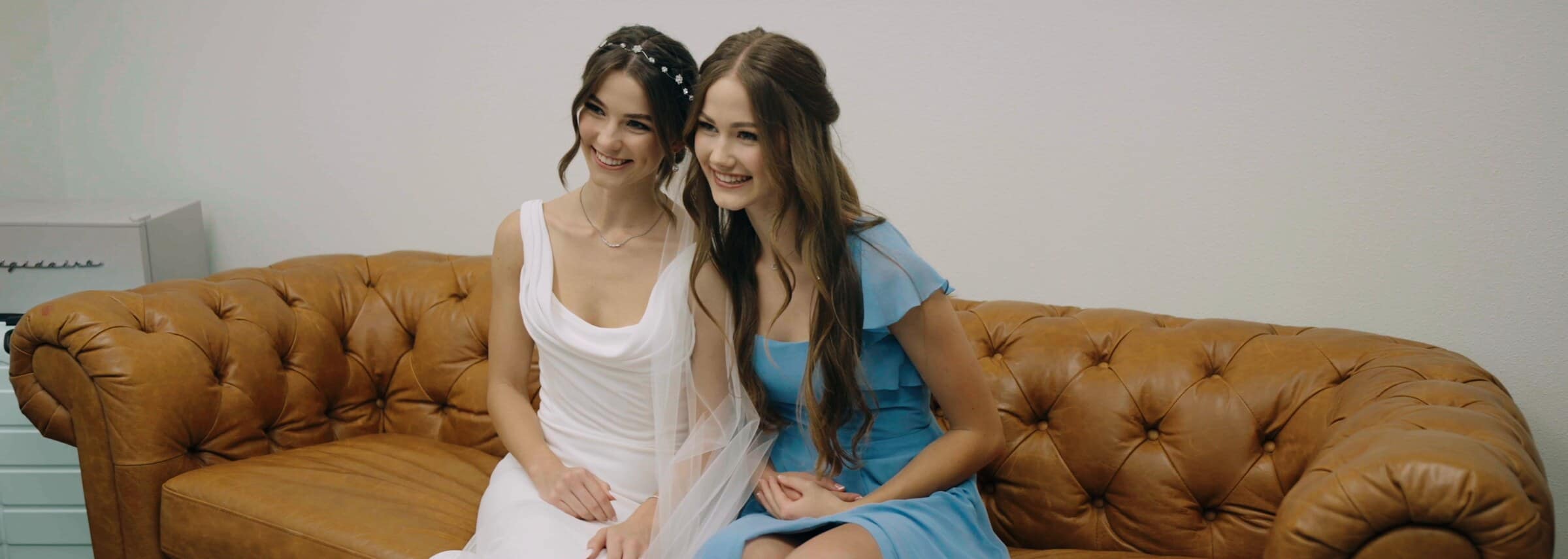 The bride laughing on a leather sofa with a friend in a blue dress