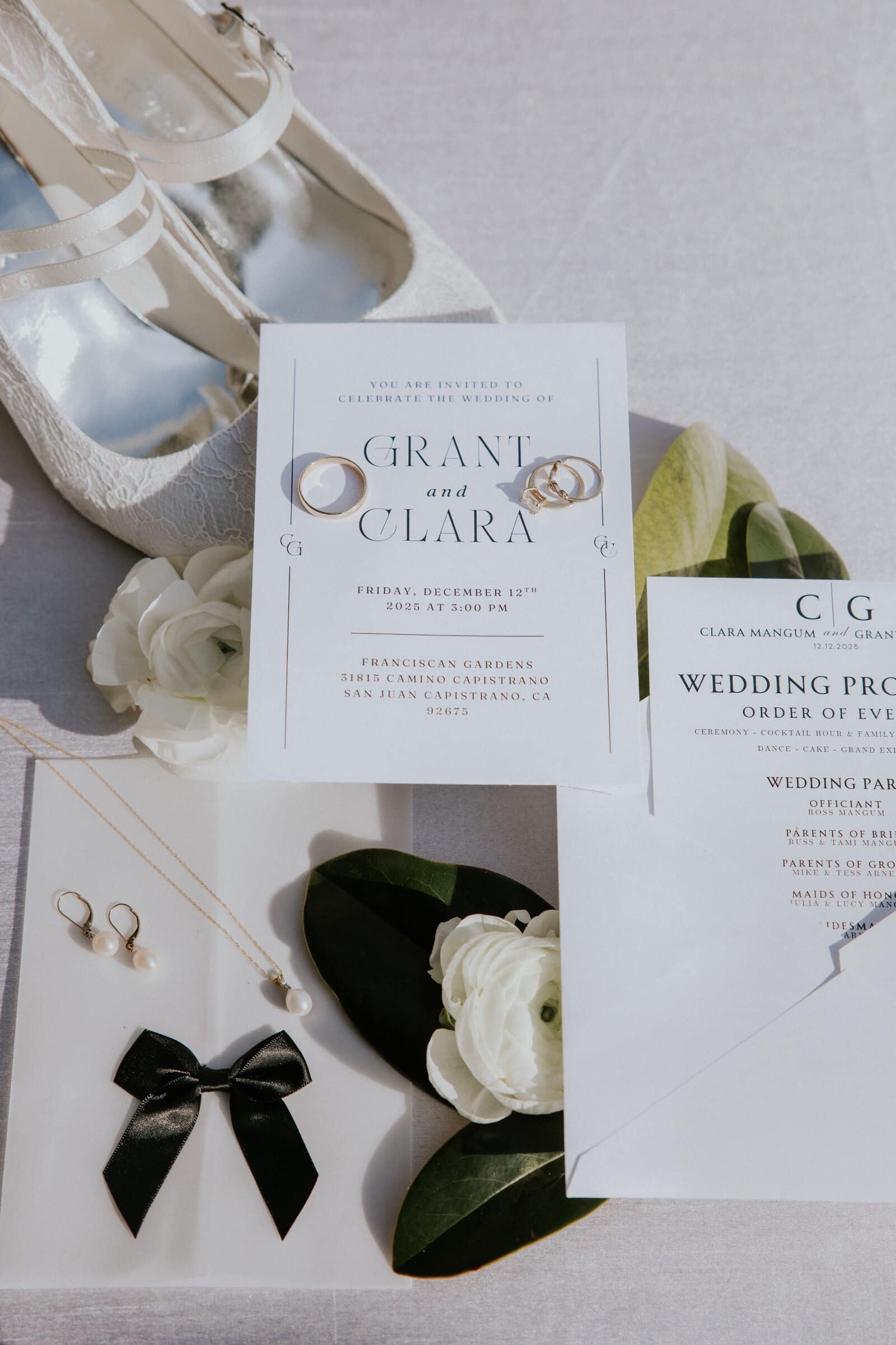 A wedding invitation for Grant and Clara with rings, a black bow tie, white roses and satin shoes