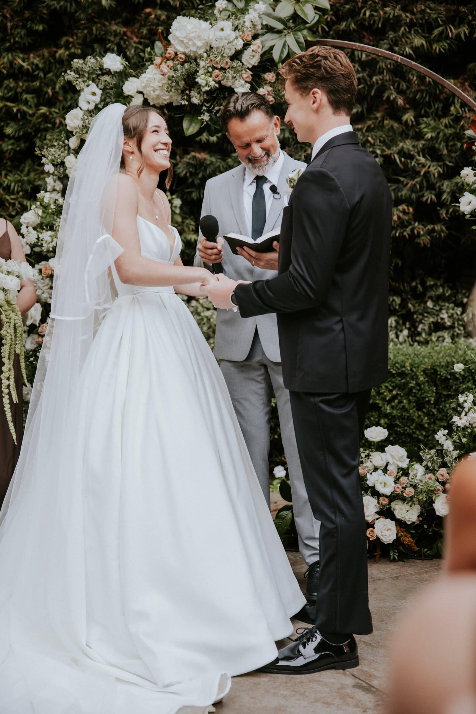 Clara and Grant facing each other at the flowered arch as the officiant speaks