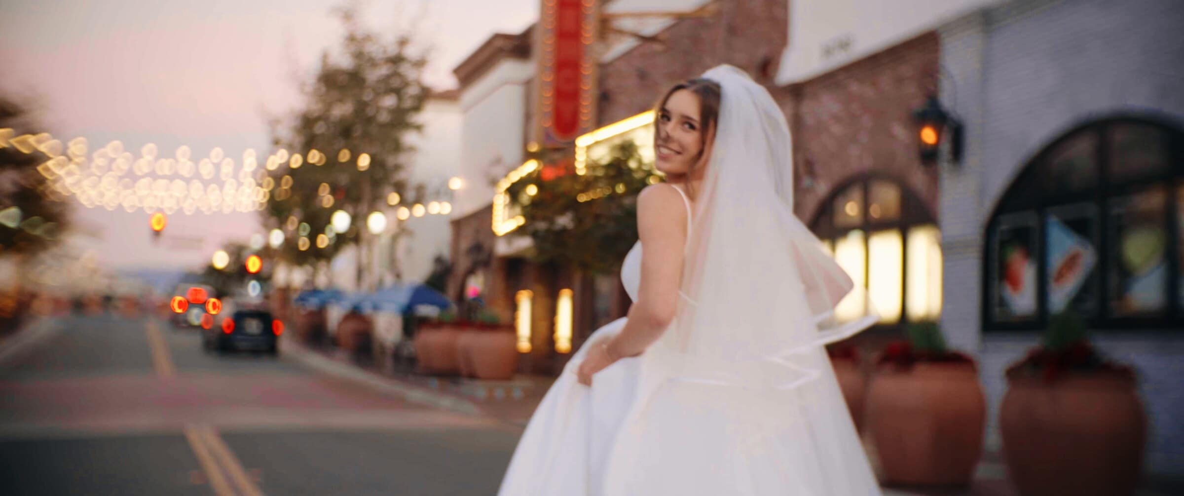 Clara in her veil on a street strung with lights at dusk