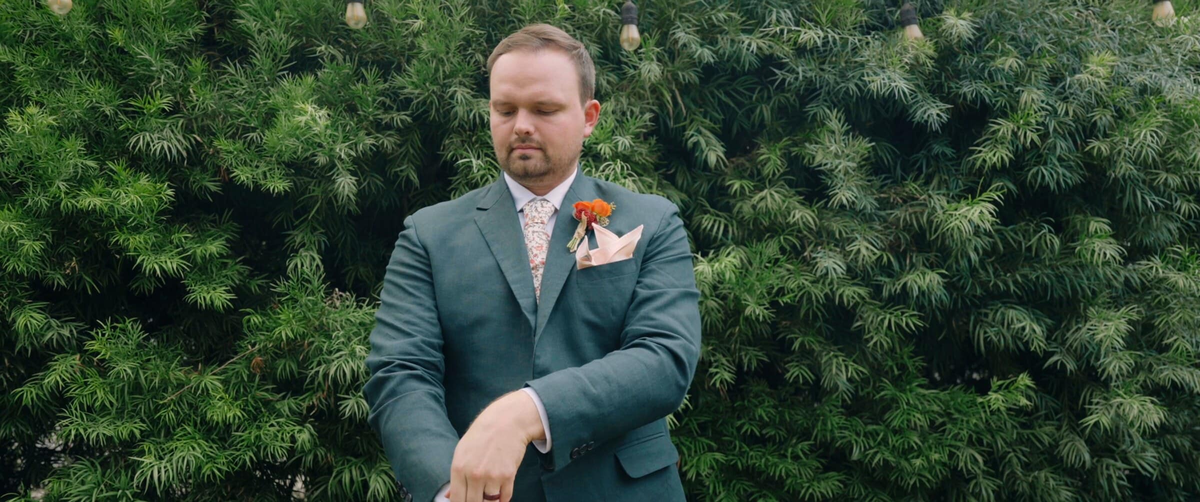 The groom in a green suit adjusting his cuff in front of a hedge