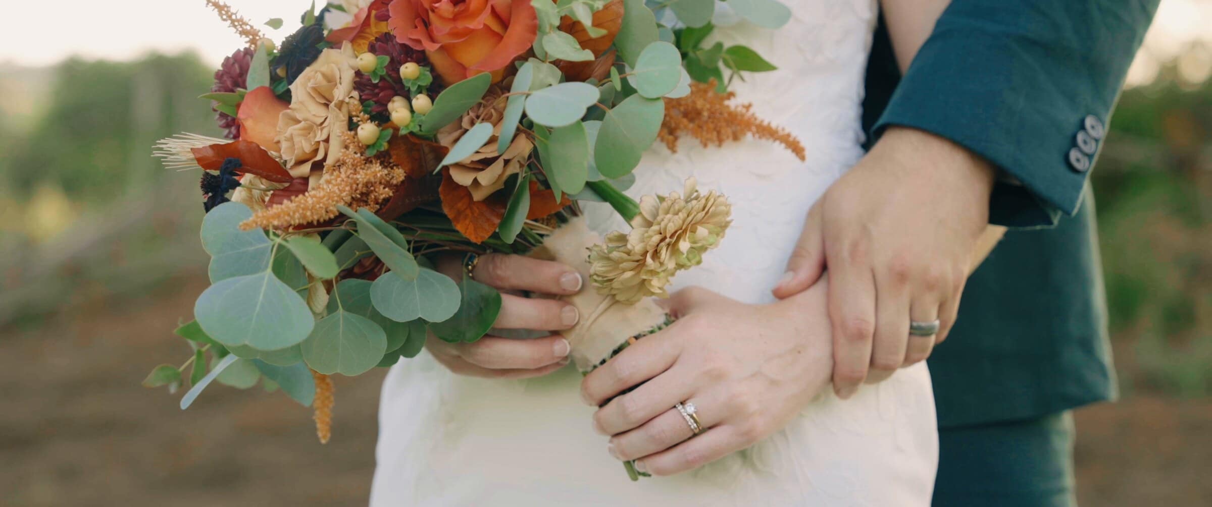 Their hands together over a bouquet of rust, peach and green, wedding rings on