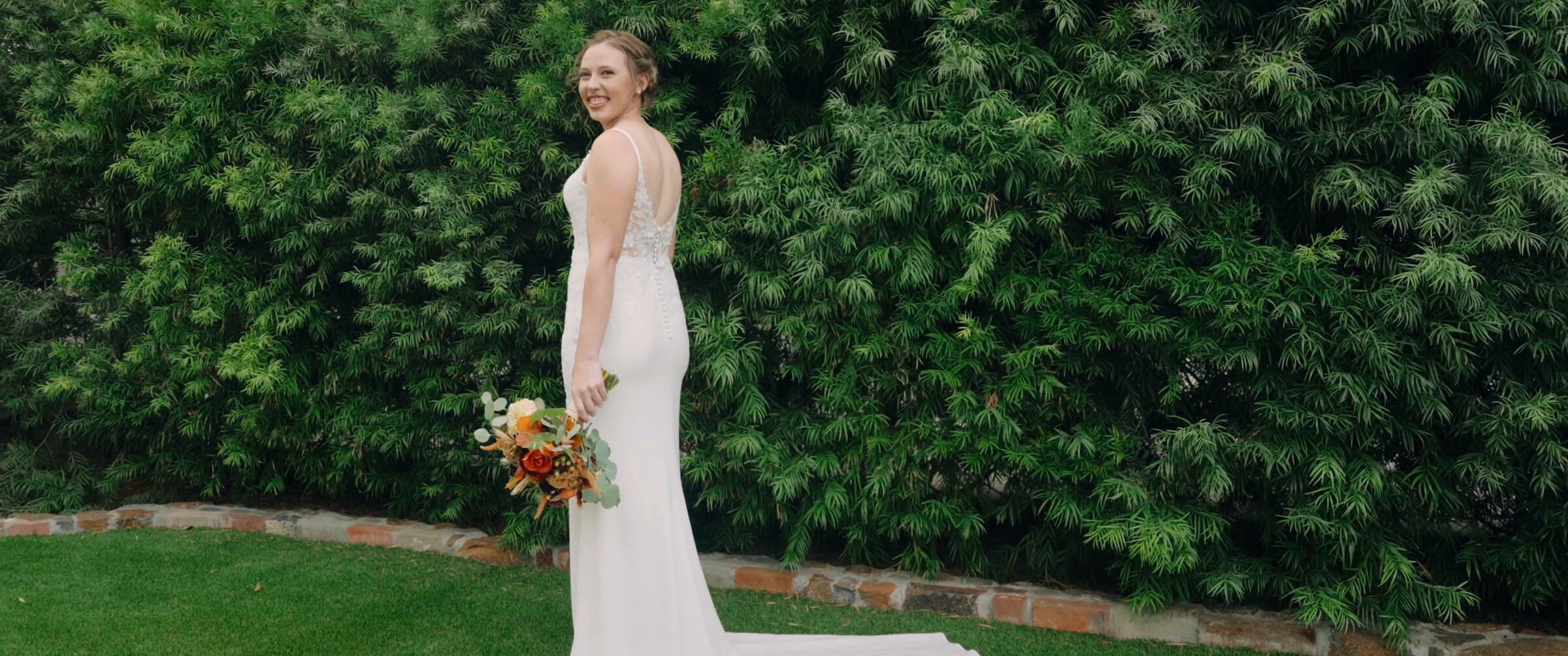 The bride looking back over her shoulder beside a tall hedge