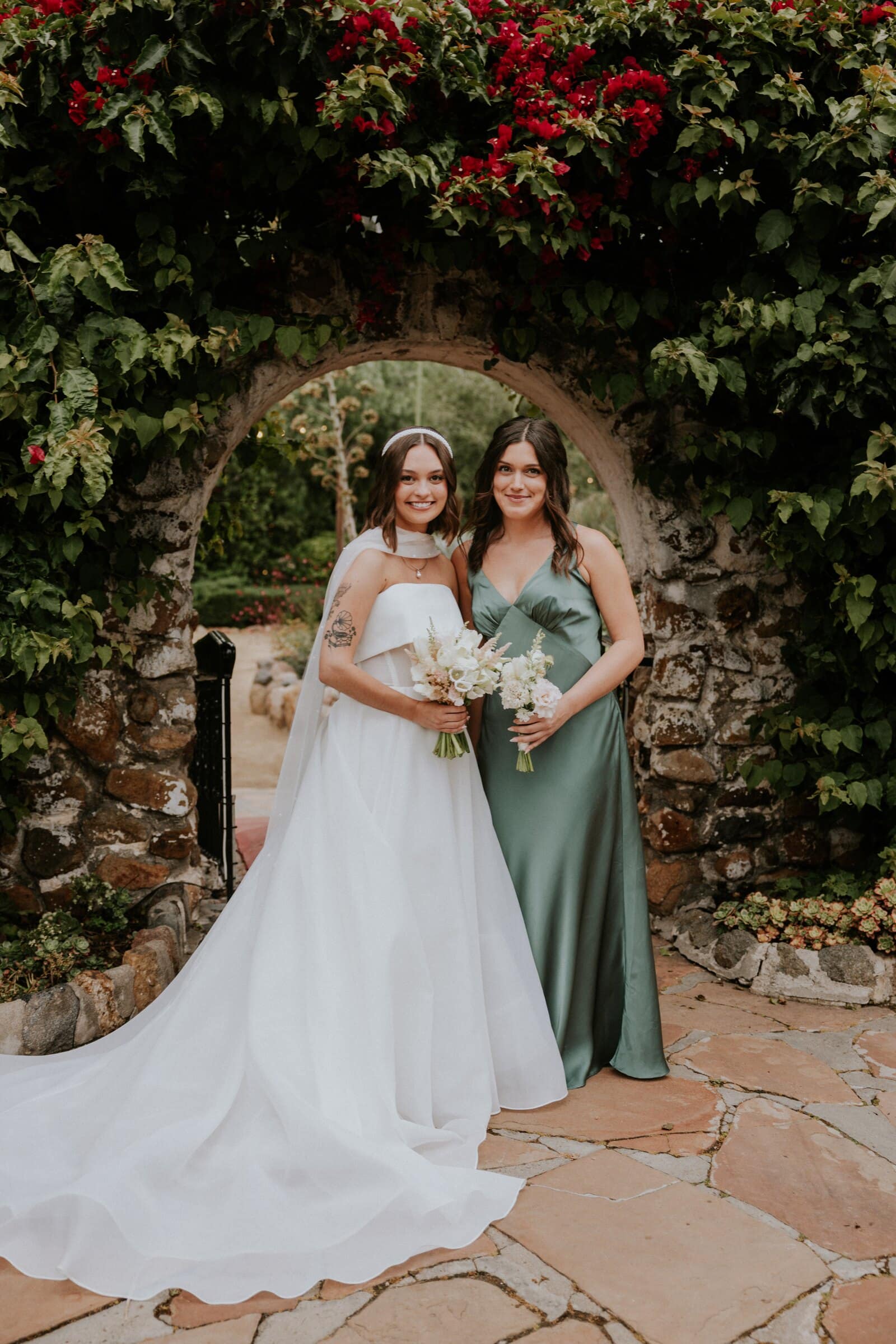 Megan with a bridesmaid in front of a stone arch and red bougainvillea