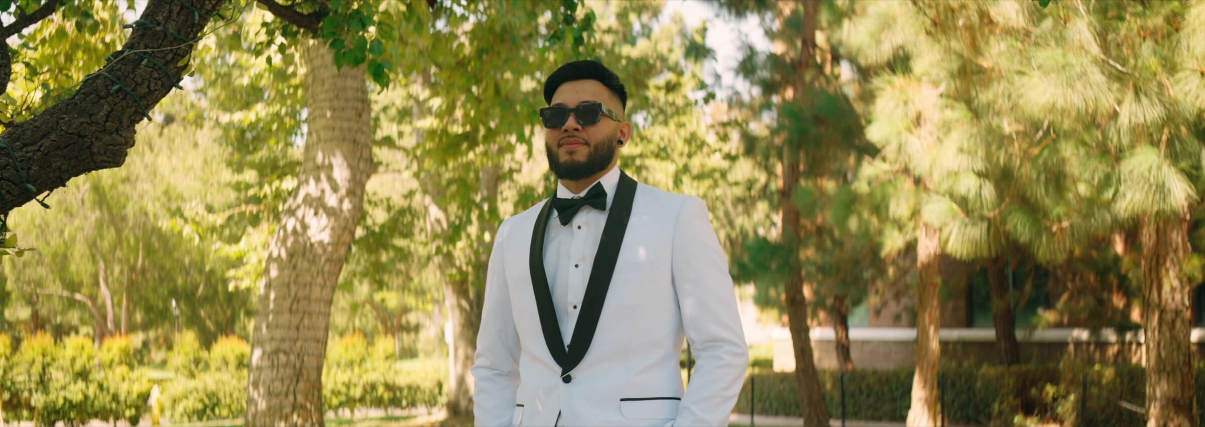 The groom in a white tuxedo and sunglasses among the trees
