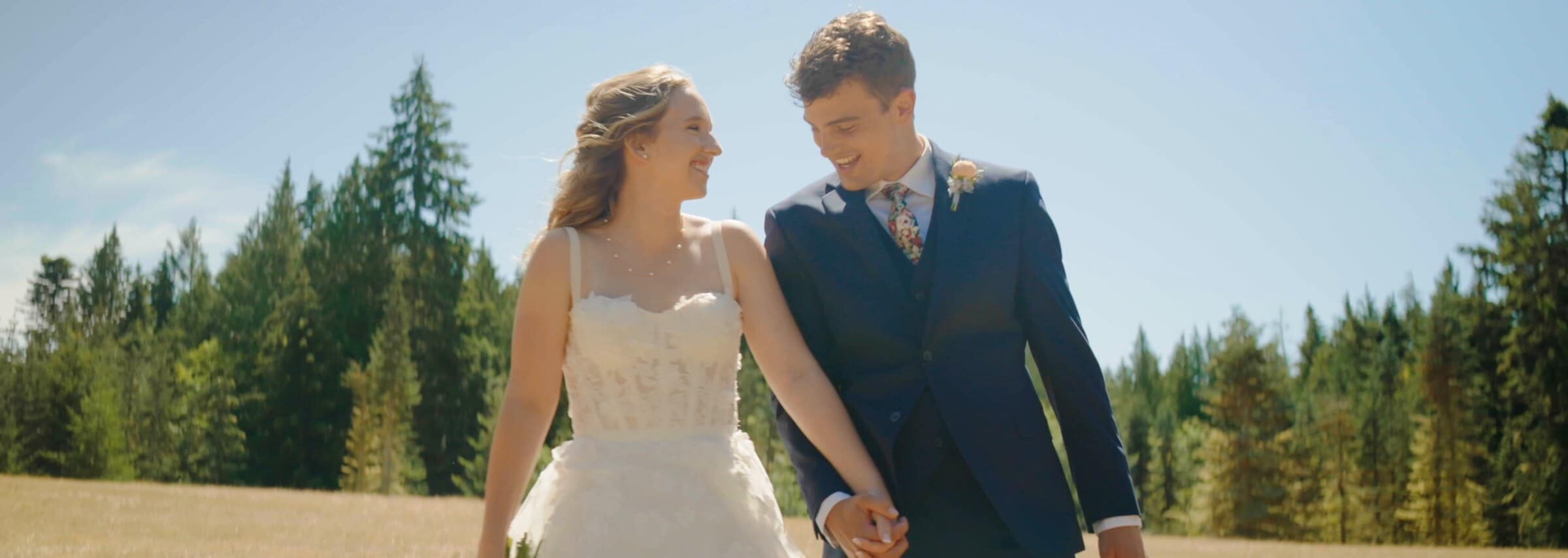 Sarah and Trent walking hand in hand through a meadow ringed by evergreens