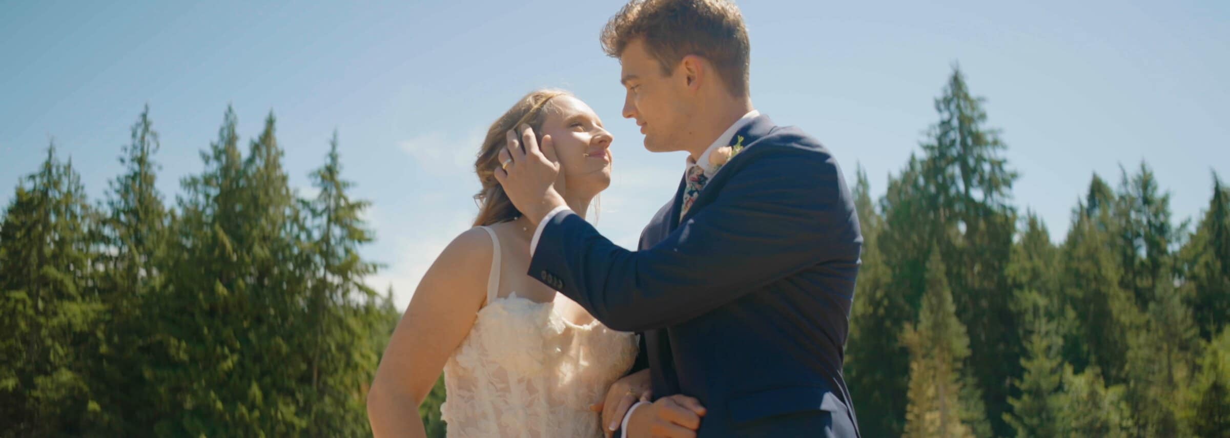 The groom touching the bride's face, tall evergreens behind them